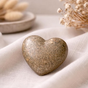May include: A heart-shaped stone with a speckled, earthy brown appearance. The stone rests on a white, textured fabric, with dried flowers and a small dish in the background. The heart is a symbol of love and affection.