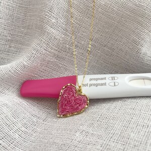 May include: A gold chain necklace with a pink heart pendant. The pendant is made of small pink pieces that are sewn together. The necklace is lying on a white fabric surface next to a pregnancy test that reads "pregnant" and "not pregnant".