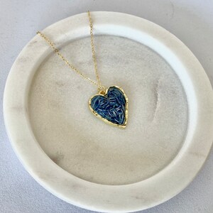 May include: A gold chain necklace with a blue heart-shaped pendant. The pendant is filled with a textured pattern of blue.