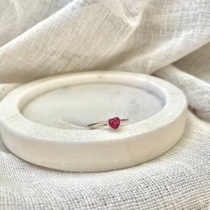 Peut inclure: Une bague en argent avec une pierre en forme de c&oelig;ur rose. La bague est pos&eacute;e sur un plat en marbre blanc.
