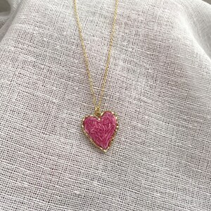May include: A gold chain necklace with a pink heart pendant. The heart is filled with small pink details, giving it a textured look.