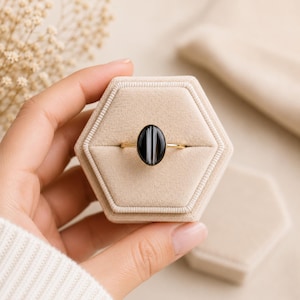 May include: A gold ring featuring an oval-shaped black stone with vertical white stripes. The ring is set into a beige velvet hexagonal ring box. The stone has a smooth, polished finish and is held by a thin, simple gold-toned metal band.