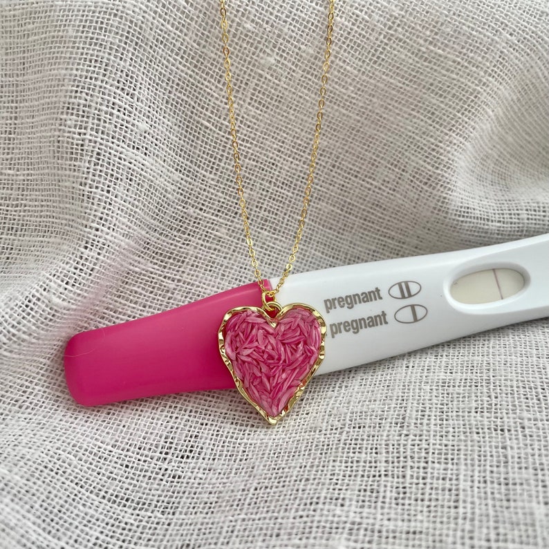 May include: A gold chain necklace with a pink heart pendant. The pendant is filled with small pink pieces. A pregnancy test with two lines is visible in the background. The test reads "pregnant" on the side.