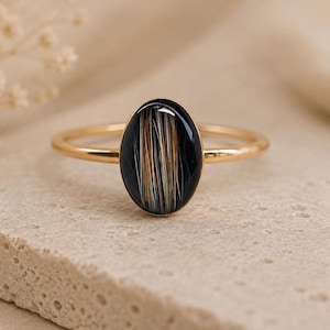 May include: A gold-toned ring featuring an oval-shaped setting with a smooth black surface. Embedded within the black oval is a vertical arrangement of fine, straight hair-like fibres in shades of white, grey, and light brown, creating a distinct pattern.