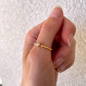 Könnte beinhalten: Ein zarter goldener Ring mit einem kleinen, runden, klaren Edelstein, der in einer Lünette gefasst ist. Der Ring hat ein dünnes Band und ein schlichtes, elegantes Design. Der Ring wird an einem Finger gezeigt.
