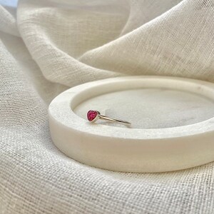 Peut inclure: Une bague en argent avec une pierre pr&eacute;cieuse en forme de c&oelig;ur rose. La bague est sur une surface en marbre blanc.