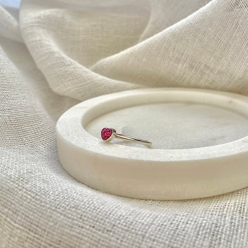 Peut inclure: Une bague en argent avec une pierre pr&eacute;cieuse en forme de c&oelig;ur rose. La bague est sur une surface en marbre blanc.