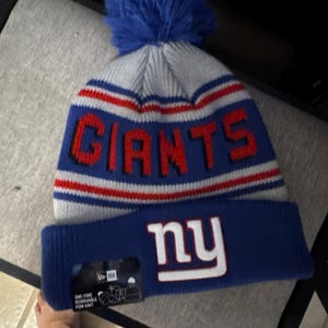 May include: A blue, white, and red knitted beanie hat with the word "GIANTS" in red block letters. The hat has a blue pom-pom on top and the letters "ny" in white on the folded brim.