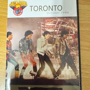 May include: A DVD case with a black background and gold trim. The front of the case features a photo of the Jackson 5 performing on stage. The text "Jacksons" is in a gold font with a red and white torch logo. The text "Toronto" is in a large gold font. The text "October 1984" is in a smaller gold font.