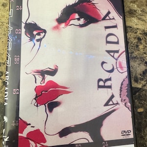 May include: DVD case with a striking illustration of a woman's face, highlighted by red lips and eye makeup. The title "ARCADIA" is visible. The case has a black border and the DVD video logo.