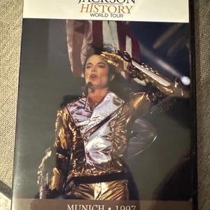 Może przedstawiać: Pudełko DVD z History World Tour Michaela Jacksona. Okładka przedstawia Michaela Jacksona na scenie w złotej i srebrnej kurtce. Tekst "MICHAEL JACKSON HISTORY WORLD TOUR" znajduje się na górze, z "MUNICH • 1997" na dole.