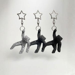 May include: Three keychains featuring abstract cat figures in gray, dark gray, and black. Each cat is attached to a silver star-shaped keyring. The keychains are displayed against a plain white background.