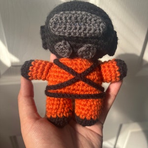 May include: A crocheted plush toy in the shape of a person wearing an orange jumpsuit with black accents. The toy has a black crocheted helmet with a gray visor.