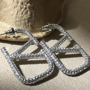 May include: A pair of silver-toned hoop earrings featuring a square-shaped design encrusted with clear, sparkling crystals. Each earring includes a post back closure and a metallic frame that is fully covered in small, reflective stones.