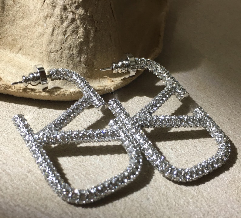 May include: A pair of silver-toned hoop earrings featuring a square-shaped design encrusted with clear, sparkling crystals. Each earring includes a post back closure and a metallic frame that is fully covered in small, reflective stones.