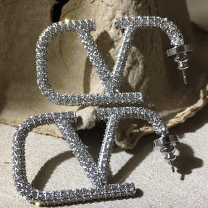 May include: A pair of silver-toned stud earrings shaped like the letters V and O, fully encrusted with small clear crystals. Each earring features a post back closure with a circular metal backing, showcasing a reflective metallic finish.