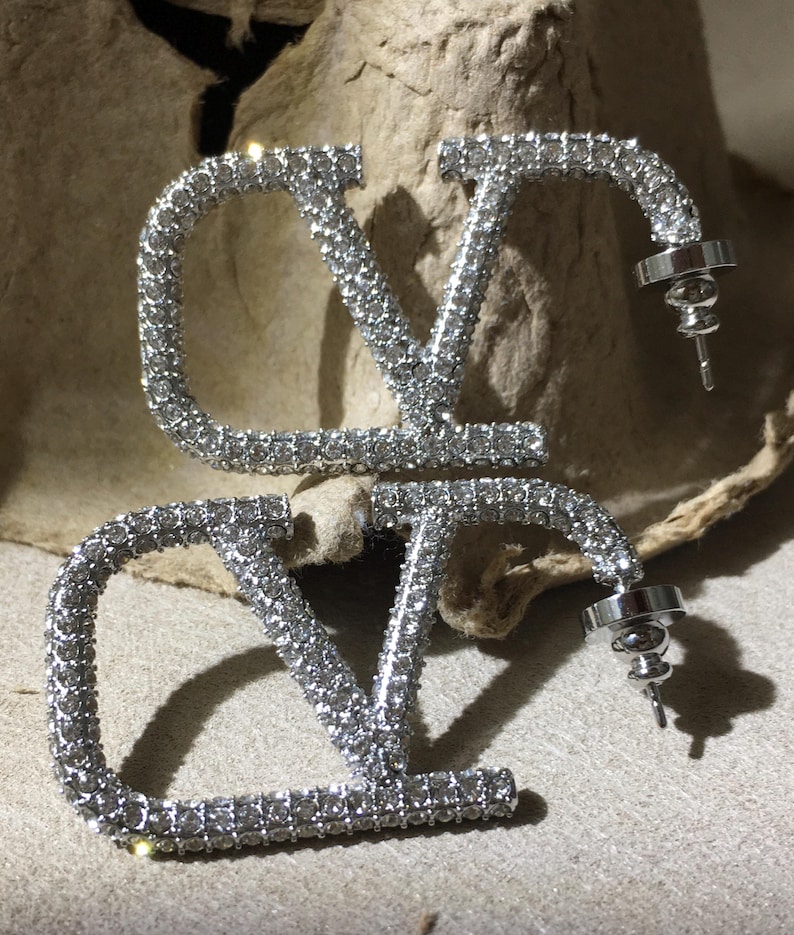May include: A pair of silver-toned stud earrings shaped like the letters V and O, fully encrusted with small clear crystals. Each earring features a post back closure with a circular metal backing, showcasing a reflective metallic finish.