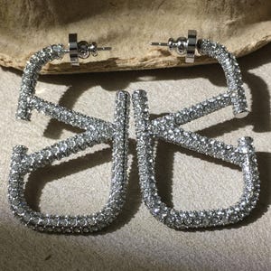 May include: A pair of silver-toned hoop earrings shaped like the letters V and K, fully encrusted with small clear rhinestones. The earrings feature a post-back closure mechanism and a metallic finish that reflects light across the stone surfaces.