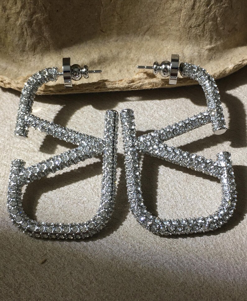 May include: A pair of silver-toned hoop earrings shaped like the letters V and K, fully encrusted with small clear rhinestones. The earrings feature a post-back closure mechanism and a metallic finish that reflects light across the stone surfaces.