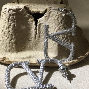 May include: A pair of silver-toned hoop earrings shaped like the letters V and L, fully encrusted with small clear crystals. Each earring features a post-back closure with a circular metal backing, showcasing a reflective, sparkling surface.