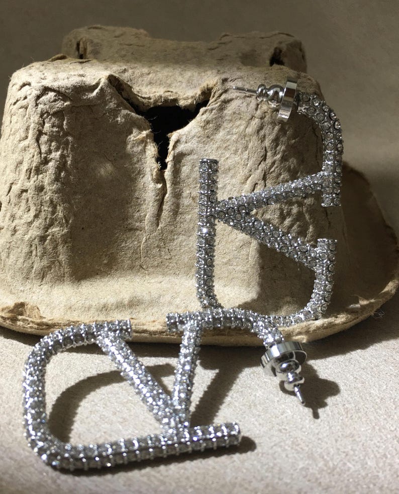 May include: A pair of silver-toned hoop earrings shaped like the letters V and L, fully encrusted with small clear crystals. Each earring features a post-back closure with a circular metal backing, showcasing a reflective, sparkling surface.