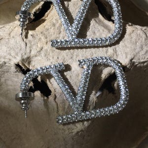 May include: A pair of silver-toned earrings featuring the letters V and D connected in a stylized design. The entire surface of the metal letters is encrusted with small, clear, reflective crystals. Each earring includes a standard post and backing.