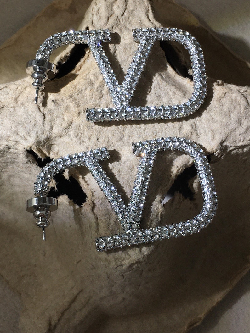 May include: A pair of silver-toned earrings featuring the letters V and D connected in a stylized design. The entire surface of the metal letters is encrusted with small, clear, reflective crystals. Each earring includes a standard post and backing.
