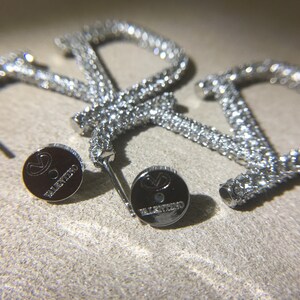 May include: A pair of silver-toned hoop earrings shaped like the letter V, encrusted with clear crystals. The earrings include two circular metal earring backs, each engraved with the Valentino logo and the brand name in small capital letters.