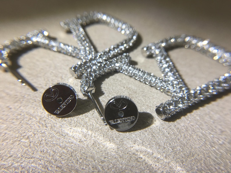 May include: A pair of silver-toned hoop earrings shaped like the letter V, encrusted with clear crystals. The earrings include two circular metal earring backs, each engraved with the Valentino logo and the brand name in small capital letters.