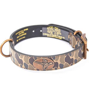 May include: A dog collar with a camouflage pattern in shades of brown, tan, and green. The collar has a bronze-colored buckle and a matching D-ring. An oval bronze emblem features a flying duck. The text "ZEP PRO" is visible.