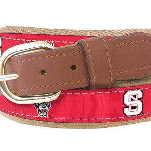 May include: A red and tan woven belt with a brown leather buckle. The belt features a repeated pattern of a white "NC" logo and a black and white mascot.