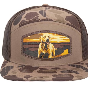 Puede incluir: Gorra de béisbol marrón y camuflaje con parte trasera de malla. La parte delantera presenta un parche rectangular con una ilustración de golden retriever sobre un fondo de puesta de sol. Adecuada para actividades al aire libre.