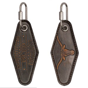 May include: A brown leather keychain with a silver hook. The keychain is shaped like a hotel key and has the text "University of Texas Est. 1883 Longhorns" on one side and a brown Longhorn steer head on the other side.