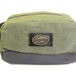 Könnte beinhalten: Eine grüne Reisetasche aus Canvas mit braunem Lederbesatz und einem braunen Alligator-Logo-Patch.