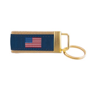 May include: A keychain featuring a blue fabric band with the American flag, stitched onto a tan leather strap. The keychain has a gold-colored metal clasp and ring.