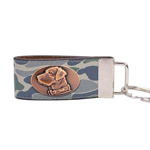 May include: A key chain with a camouflage pattern and a bronze-colored oval emblem featuring a dog's head. The key chain has a silver-colored metal loop for attaching keys.