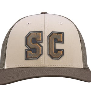 May include: A beige and brown baseball cap featuring a mesh back and a solid front panel. The front displays the letters SC in a dark, weathered, block-style font with a stitched border, centred on the beige fabric above the brown brim.