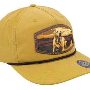 May include: Mustard yellow baseball cap with a black rope detail and a curved brim. The cap features a rectangular patch with a dog illustration set against a sunset backdrop. A small circular emblem is on the brim.