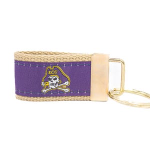Peut inclure: Un porte-clés violet et beige avec un fermoir et un anneau en métal doré. La bande violette présente le logo ECU Pirates en or, blanc et jaune. Le porte-clés est en tissu tissé.