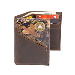 May include: Brown leather wallet with a camouflage accent and a decorative metal button. The wallet is open, revealing card slots and cash. The accent features a leaf pattern and the text "12 GA".