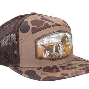 May include: A brown and tan camouflage baseball cap with a dark brown mesh back and a flat brim. The front panel features a rectangular leather-trimmed patch displaying a detailed illustration of a brown and white hunting dog in a field.