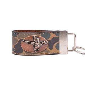 May include: A leather keychain with a camouflage pattern. It features a bronze-coloured oval emblem with a flying duck design. The keychain has a silver-coloured metal clasp and a key ring.