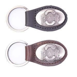 May include: Two keychains with the Ohio State logo. One keychain has a black leather strap, and the other has a brown leather strap. Both have a silver-colored oval emblem with the words "OHIO STATE" in a stylized font.