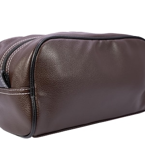 Puede incluir: Un neceser de cuero marr&oacute;n con superficie texturizada. El neceser tiene ribete negro, cremallera cosida en blanco y un peque&ntilde;o asa negra en el lateral. El neceser es rectangular y parece vac&iacute;o.