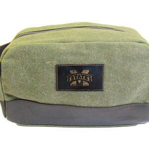 Peut inclure: Une trousse de toilette en toile verte avec une bordure en cuir marron et un patch en cuir marron avec le texte "M STATE" en lettres dorées.