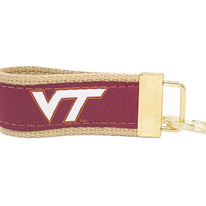 May include: A maroon and tan keychain with a gold-colored metal clasp and key rings. The maroon section features the white letters "VT" in a stylized font. The keychain is designed with a woven tan strap.