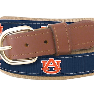 May include: A brown leather belt with a navy blue and orange woven strap featuring the Auburn University logo. The belt has a gold buckle.