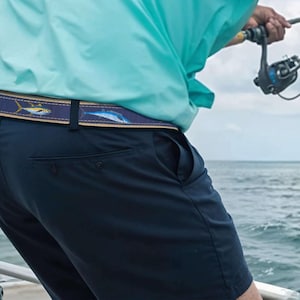 May include: A navy blue belt with a repeating fish pattern in blue and yellow. The belt is worn with navy shorts and a light blue shirt. The person is fishing on a boat, with the ocean in the background.