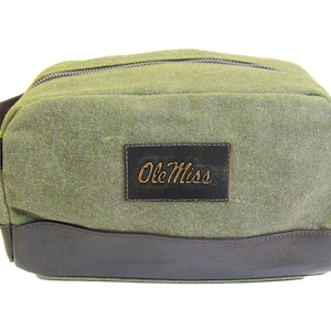 Könnte beinhalten: Eine grüne Toilettentasche aus Canvas mit braunem Lederbesatz und einem braunen Lederflicken mit dem Text "Ole Miss" in goldenen Buchstaben.