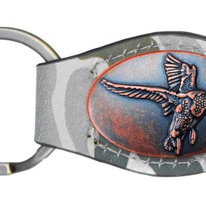 May include: A camouflage leather keychain with a metal oval charm featuring a copper-colored duck in flight.
